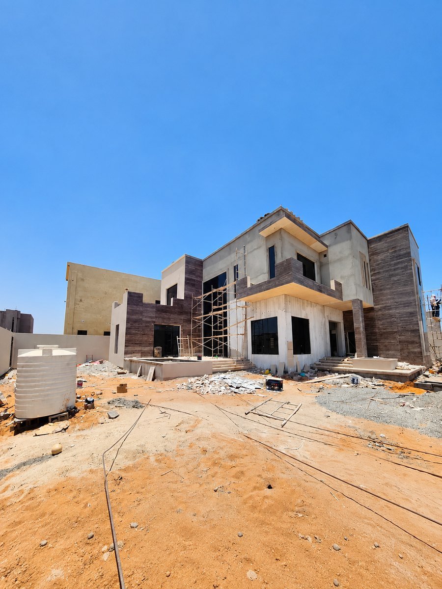 Site photograph of a two-storey villa under construction, with scaffolding on the façade and water tanks on the roof